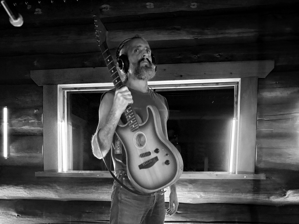 Achille Alyster debout dans le studio de La Renégate, casque sur les oreilles, guitare à l'épaule, en noir et blanc
