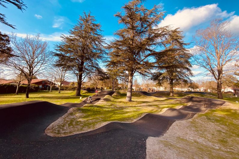 Un pump track en enrobé noir aux bosses arrondies, entre la pelouse et de grands arbres